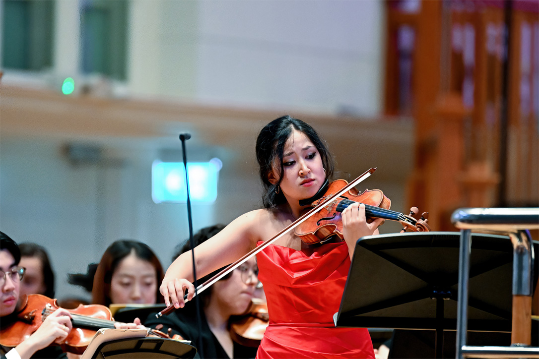 A women wearing a red dress, playing the violin next to the conductor's podium, with an orchestra behind them.